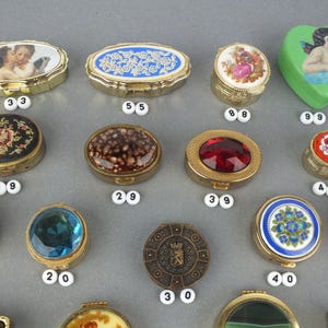 Op de afbeelding: Een verzameling decoratieve snuisterijendoosjes in verschillende vormen, waaronder ovaal, rond en hartvormig. De doosjes hebben sierlijke ontwerpen, zoals cherubs, bloempatronen en juweelachtige accenten. De doosjes zijn gemaakt van metaal en hebben scharnierende deksels.
