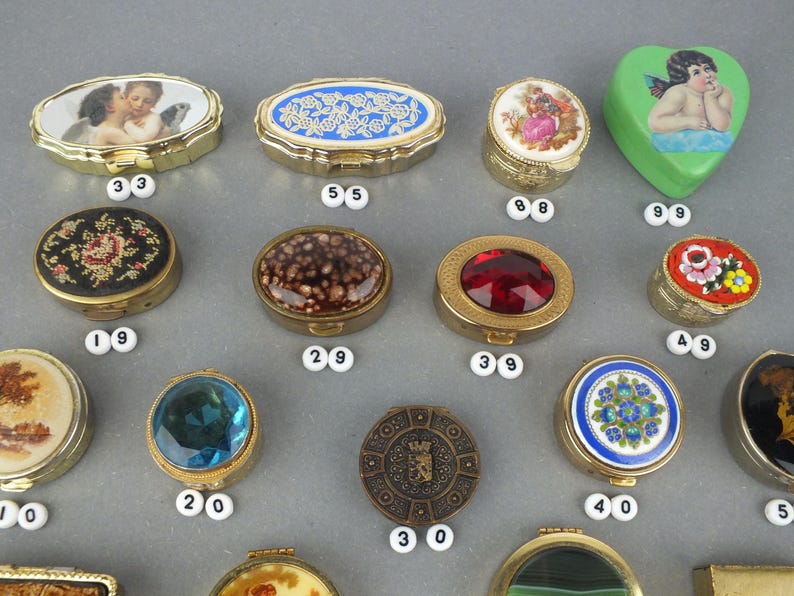 Op de afbeelding: Een verzameling decoratieve snuisterijendoosjes in verschillende vormen, waaronder ovaal, rond en hartvormig. De doosjes hebben sierlijke ontwerpen, zoals cherubs, bloempatronen en juweelachtige accenten. De doosjes zijn gemaakt van metaal en hebben scharnierende deksels.