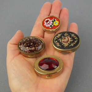 Op de afbeelding: Vier kleine, antieke pillendoosjes in een hand. De doosjes zijn ovaal van vorm met goudkleurige metalen frames. E&eacute;n heeft een rood moza&iuml;ek bloemenontwerp, een ander een donkerbruine gespikkelde bovenkant, een derde een rode edelsteen en de laatste een bloemenborduurontwerp.