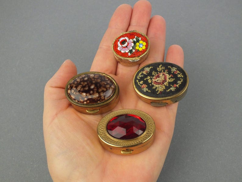 Op de afbeelding: Vier kleine, antieke pillendoosjes in een hand. De doosjes zijn ovaal van vorm met goudkleurige metalen frames. E&eacute;n heeft een rood moza&iuml;ek bloemenontwerp, een ander een donkerbruine gespikkelde bovenkant, een derde een rode edelsteen en de laatste een bloemenborduurontwerp.