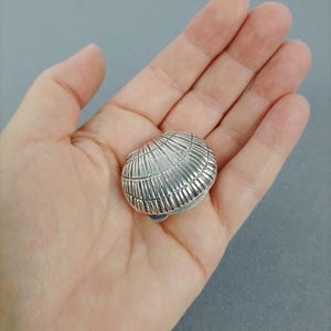 Könnte beinhalten: Eine kleine, glänzend silberfarbene Metallschmuckdose in Form einer Muschel. Die Außenseite weist eine detaillierte, gerippte Textur auf, die die natürlichen Rillen einer Muschel nachahmt, mit einem seitlich sichtbaren Scharniermechanismus.