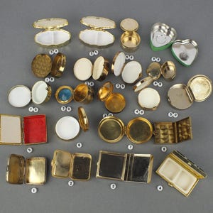 Op de afbeelding: Een verzameling kleine, vintage metalen compacts en containers in verschillende vormen, waaronder rond, ovaal en hartvormig. De compacts zijn goudkleurig, sommige met witte, rode en blauwe accenten. Sommige zijn open en onthullen spiegels of compartimenten.