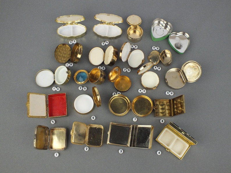 Op de afbeelding: Een verzameling kleine, vintage metalen compacts en containers in verschillende vormen, waaronder rond, ovaal en hartvormig. De compacts zijn goudkleurig, sommige met witte, rode en blauwe accenten. Sommige zijn open en onthullen spiegels of compartimenten.