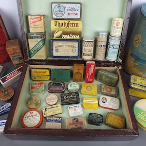 Könnte beinhalten: Eine Sammlung von alten Metalldosen und Behältern, darunter eine große hölzerne Präsentationsbox, gefüllt mit verschiedenen kleinen rechteckigen und runden Arznei- und Lebensmitteldosen mit farbigen Etiketten, Texten und Grafiken, sowie einigen losen Dosen.