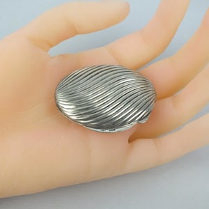 Könnte beinhalten: Eine kleine, ovale Pillendose aus Silber mit einer strukturierten Oberfläche, die durch diagonale, erhabene Rippen an eine Muschel erinnert. Das metallische Objekt hat eine polierte Oberfläche und einen Klappdeckel mit einem Muster.