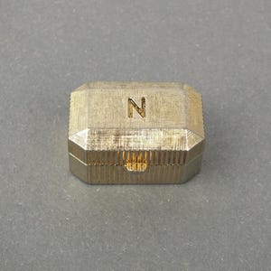 Könnte beinhalten: Eine goldfarbene, geometrische Schmuckschatulle mit strukturierter Oberfläche. Die Schatulle hat eine achteckige Form mit einem Klappdeckel und einem kleinen Verschluss. Der Buchstabe "N" ist auf der Oberseite eingraviert.