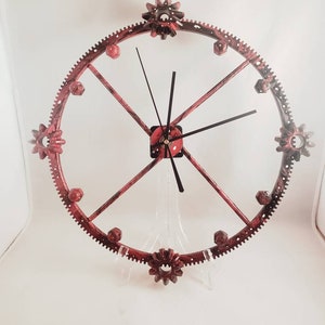May include: A red and black metal wall clock with a gear design. The clock has a circular frame with gear-shaped accents and black clock hands. The clock is supported by a clear stand against a white background.