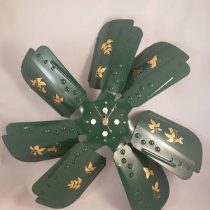 May include: A green metal fan blade clock with gold leaf accents. The clock has a white face with black hands and a white background.