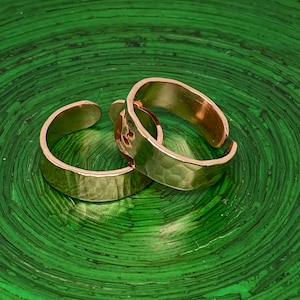 May include: Two hammered gold rings on a green background. The rings are simple and have a textured finish.