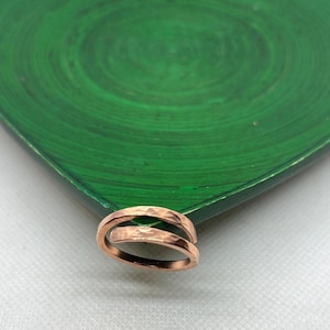 May include: A copper-coloured bypass ring featuring a hammered, textured surface. The band is open at the front, with two overlapping ends that create a continuous, layered appearance. The metal has a warm, metallic finish with visible indentations.