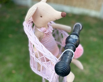 Puede incluir: Un asiento de muñeca de mimbre tejido en color rosa claro sujeto al manillar negro de un patinete. Una muñeca de tela beige con nariz marrón y un conjunto de lunares rosas con cuello de volantes se sienta en el asiento, que tiene un enganche blanco.
