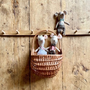 Pode incluir: Duas bonecas de rato branco vestidas estão sentadas em uma cesta de vime marrom pendurada em uma parede de madeira com ganchos. Uma terceira boneca de rato está pendurada em um gancho acima da cesta.