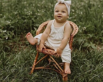 Pode incluir: Uma menina pequena vestindo um vestido branco e uma faixa de cabeça rosa claro está sentada em uma cadeira de vime. A cadeira está em um campo de grama verde.