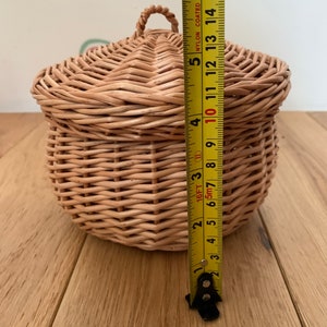 Op de afbeelding: Een ronde, geweven rieten mand met een deksel en een handvat. De mand is ongeveer 13 cm hoog. De natuurlijk gekleurde mand staat op een houten oppervlak. Het deksel heeft een afgeronde vorm.