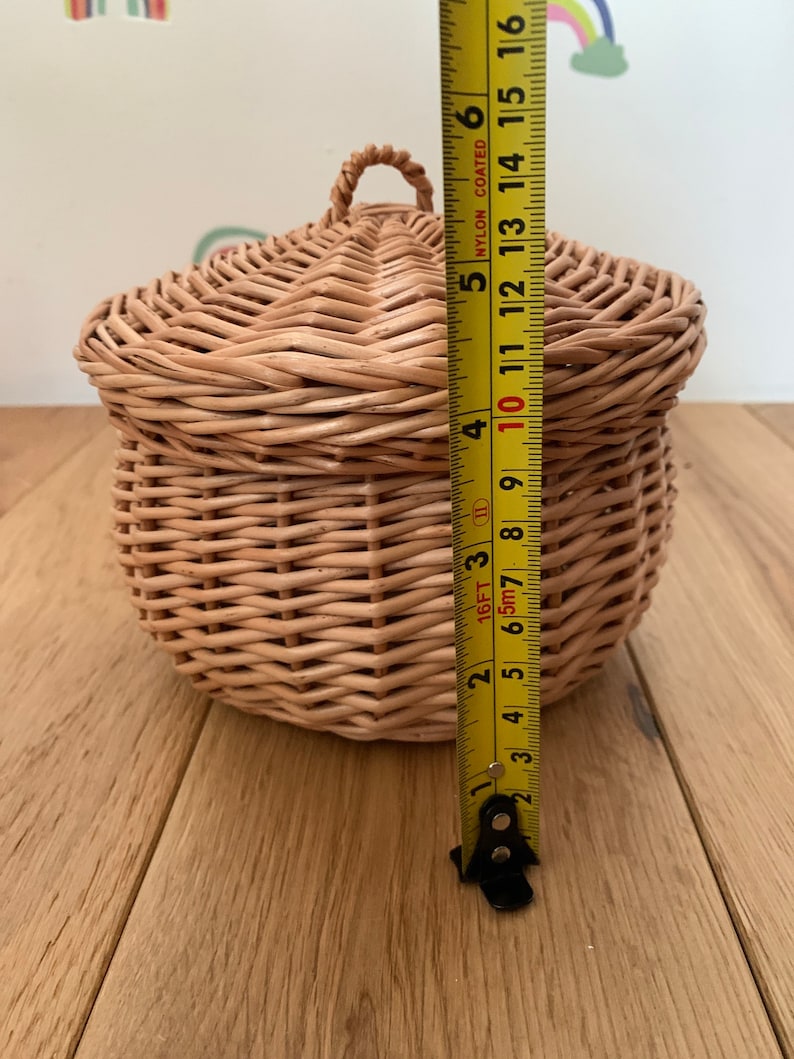 Op de afbeelding: Een ronde, geweven rieten mand met een deksel en een handvat. De mand is ongeveer 13 cm hoog. De natuurlijk gekleurde mand staat op een houten oppervlak. Het deksel heeft een afgeronde vorm.