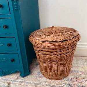 May include: A round, light brown woven wicker basket with a matching lid featuring a small central handle. The basket has a vertical weave pattern and is positioned next to a teal-colored chest of drawers with black knobs.