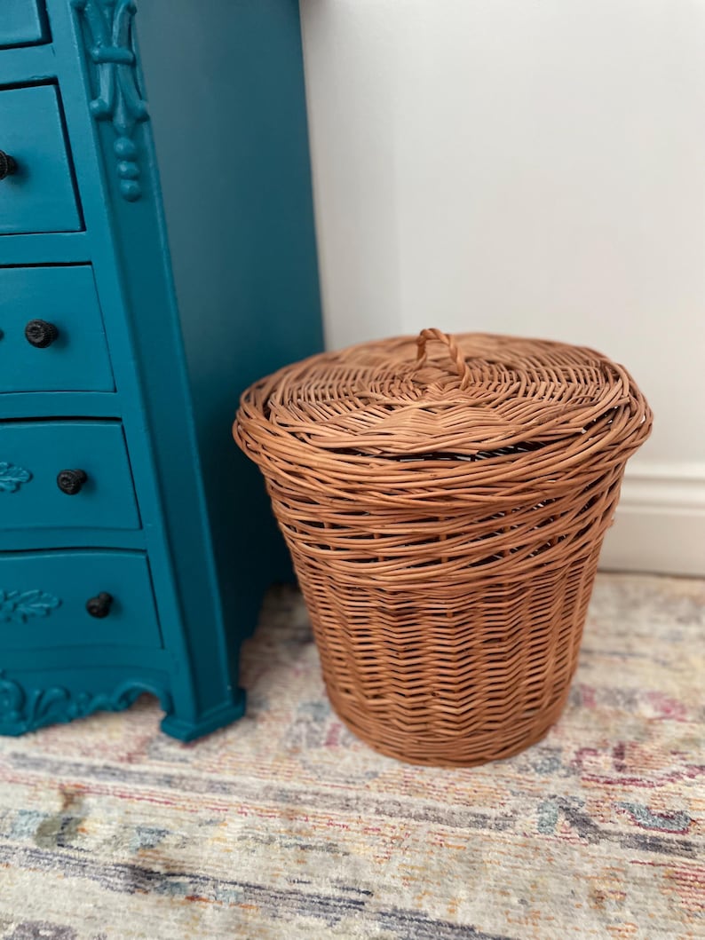 May include: A round, light brown woven wicker basket with a matching lid featuring a small central handle. The basket has a vertical weave pattern and is positioned next to a teal-colored chest of drawers with black knobs.