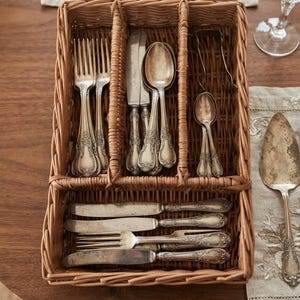 May include: A rectangular woven wicker cutlery organizer with three vertical compartments and one horizontal compartment. The organizer contains various silver-toned metal forks, knives, and spoons with ornate embossed handles.