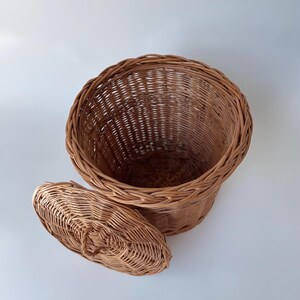 May include: A round, light brown woven wicker basket with a matching circular lid. The basket features a deep, open interior and a braided rim design. The lid is detached and rests beside the basket, showing a woven pattern and a small handle.