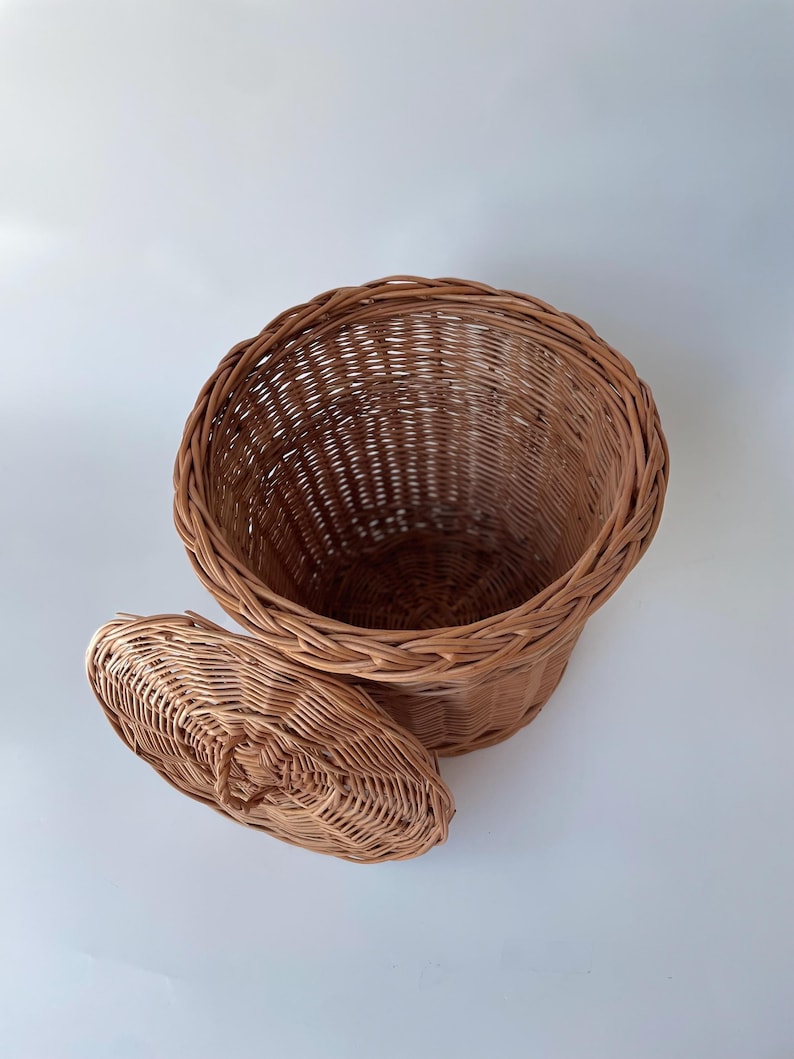 May include: A round, light brown woven wicker basket with a matching circular lid. The basket features a deep, open interior and a braided rim design. The lid is detached and rests beside the basket, showing a woven pattern and a small handle.