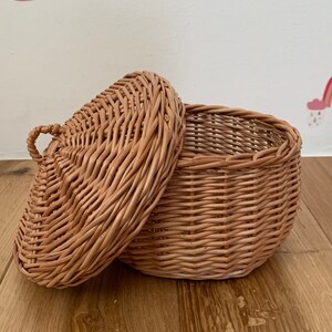 Op de afbeelding: Een kleine, ronde, geweven rieten mand met een deksel. De mand is lichtbruin en heeft een handvat op het deksel. Het deksel is gedeeltelijk open en onthult de binnenkant. De mand staat op een houten oppervlak.