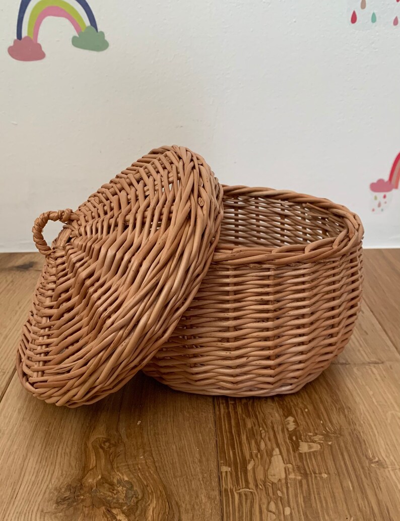 Op de afbeelding: Een kleine, ronde, geweven rieten mand met een deksel. De mand is lichtbruin en heeft een handvat op het deksel. Het deksel is gedeeltelijk open en onthult de binnenkant. De mand staat op een houten oppervlak.
