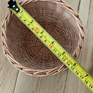 Op de afbeelding: Een ronde, geweven rieten mand met een gele meetlint over de bovenkant. De mand is lichtbruin en lijkt ongeveer 18 cm in diameter te zijn. De meetlint toont metingen in inches.