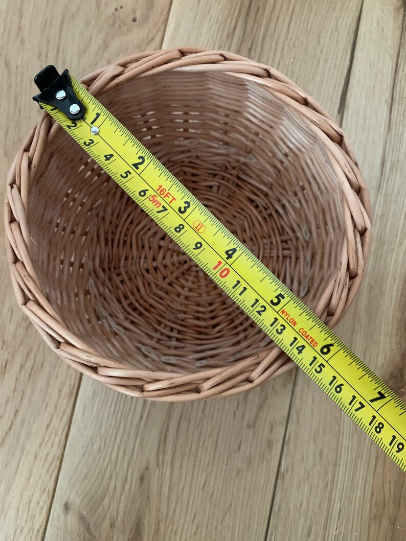 Op de afbeelding: Een ronde, geweven rieten mand met een gele meetlint over de bovenkant. De mand is lichtbruin en lijkt ongeveer 18 cm in diameter te zijn. De meetlint toont metingen in inches.
