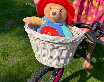 Puede incluir: Una cesta de mimbre blanca unida a una bicicleta rosa con un oso de peluche que lleva un sombrero rojo dentro. El oso lleva una camisa azul y amarilla con un corazón rojo. La bicicleta está en un césped verde.