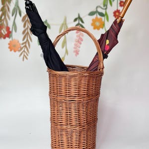 May include: A tall, woven wicker umbrella stand holds two umbrellas. One umbrella is black, and the other is burgundy with a geometric pattern. The stand has a handle and is made of light brown wicker.