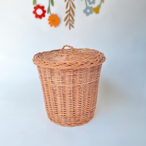 May include: A round, light brown woven wicker basket with a matching lid featuring a small, braided handle at the center. The basket has a cylindrical shape with a textured, interlaced pattern throughout the body and a reinforced rim.