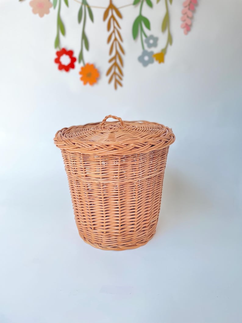 May include: A round, light brown woven wicker basket with a matching lid featuring a small, braided handle at the center. The basket has a cylindrical shape with a textured, interlaced pattern throughout the body and a reinforced rim.