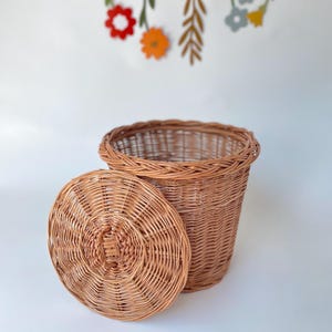 May include: A round, light brown woven wicker basket with a matching circular lid resting beside it. The basket features a sturdy, braided rim and a tight, uniform weave pattern throughout the body and the lid, showcasing natural wood tones.