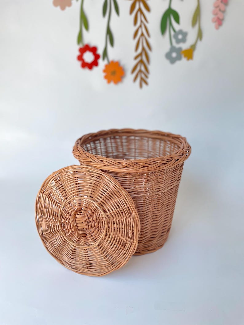 May include: A round, light brown woven wicker basket with a matching circular lid resting beside it. The basket features a sturdy, braided rim and a tight, uniform weave pattern throughout the body and the lid, showcasing natural wood tones.