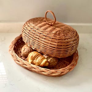 Op de afbeelding: Een geweven rieten broodmand met een deksel en een handvat. De mand is gevuld met broodjes en een croissant.