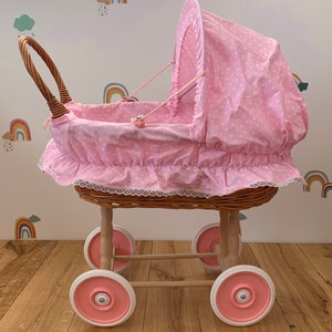 May include: A traditional doll's pram with a pink and white polka dot hood and interior. The pram has a woven wicker basket, wooden frame, and pink wheels. The hood is adjustable, and the pram has a classic design, reminiscent of a bygone era.