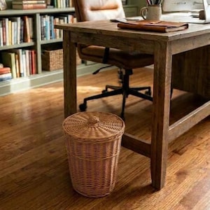 May include: A cylindrical woven wicker wastebasket with a matching lid, featuring a natural light brown finish and a tapered design, positioned on a wooden floor beneath a wooden desk in a home office setting.