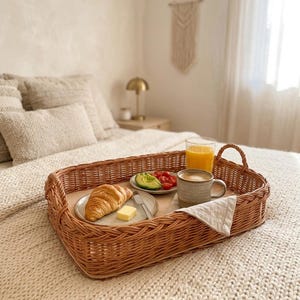 May include: A rectangular woven wicker tray with two side handles, containing a ceramic plate with a croissant and a square of butter, a small plate with sliced avocado and cherry tomatoes, a ceramic mug of coffee, and a glass of orange juice.