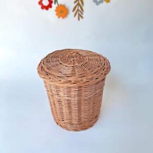 May include: A round, light brown wicker basket with a matching lid. The basket is woven with a tight pattern and has a cylindrical shape. The lid fits snugly on top. Decorative floral cutouts hang in the background.