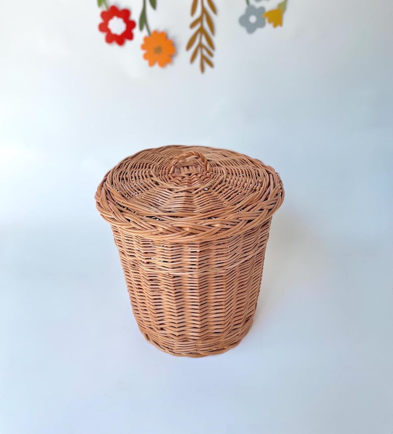 May include: A round, light brown woven wicker basket with a matching lid featuring a small central handle. The basket has a tapered cylindrical shape with a tightly woven texture, showcasing the natural tan color of the reed material.