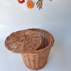 May include: A round, light brown woven wicker basket with a matching lid featuring a small central handle. The basket has a vertical ribbed texture and is positioned against a plain white background with colorful paper flower cutouts.