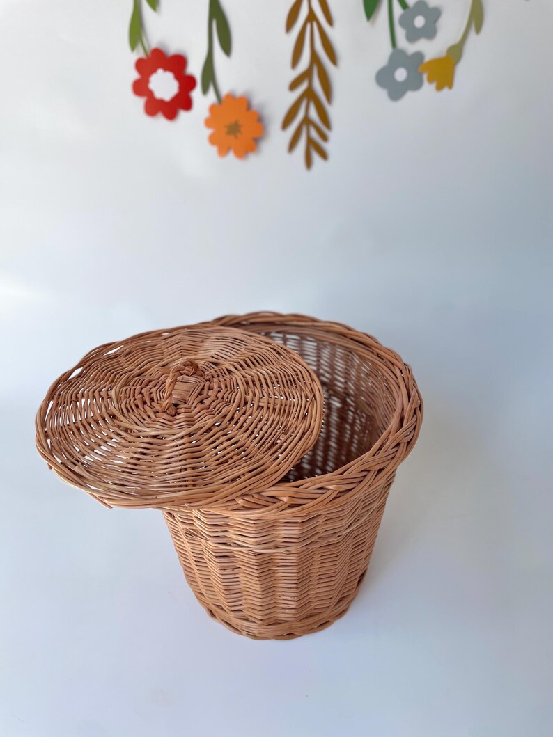May include: A round, light brown woven wicker basket with a matching lid featuring a small central handle. The basket has a vertical ribbed texture and is positioned against a plain white background with colorful paper flower cutouts.