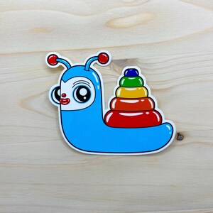 May include: A blue snail with a rainbow stacking ring on its back. The snail has a white face with red lips and large black eyes.