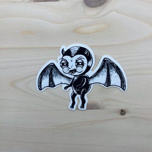 May include: A black and white sticker featuring a cartoon bat with a mischievous expression. The bat has large wings, a small body, and a devilish grin. The sticker is placed on a light-colored wooden surface.
