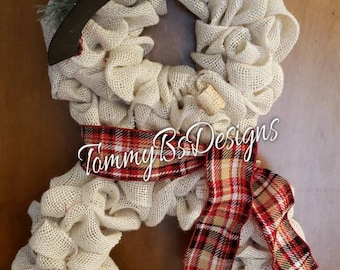May include: A burlap wreath shaped like a snowman with a black top hat and a red and black plaid bow. The wreath is decorated with pine sprigs and red berries. The text "Tommy B's Designs" is visible on the wreath.