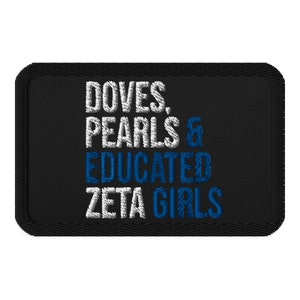 May include: Black embroidered patch with white and blue text. The text reads "DOVES, PEARLS & EDUCATED ZETA GIRLS". The patch has a black border and is rectangular in shape. The text is in a stylized font.