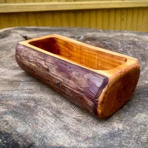 May include: A handcrafted wooden planter box with a rectangular shape. The exterior features a natural bark texture with shades of brown and purple, while the interior is a lighter, polished wood. The planter is designed to hold small plants or succulents.