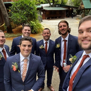 May include: Six men in navy blue suits with rust-colored ties and boutonnieres stand in a group. They are smiling and looking at the camera. The men are in a courtyard setting with a building in the background.