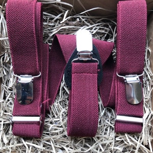 May include: Three burgundy suspenders with silver metal clips. The suspenders are made of a textured fabric and are arranged in a box filled with brown packing material.