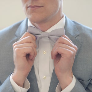 May include: A man adjusts a light gray bow tie. The bow tie is made of a soft, textured fabric.